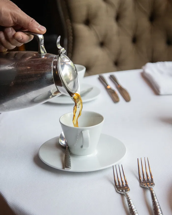 Mano sirviendo café desde una cafetera plateada en una taza blanca sobre una mesa de restaurante.